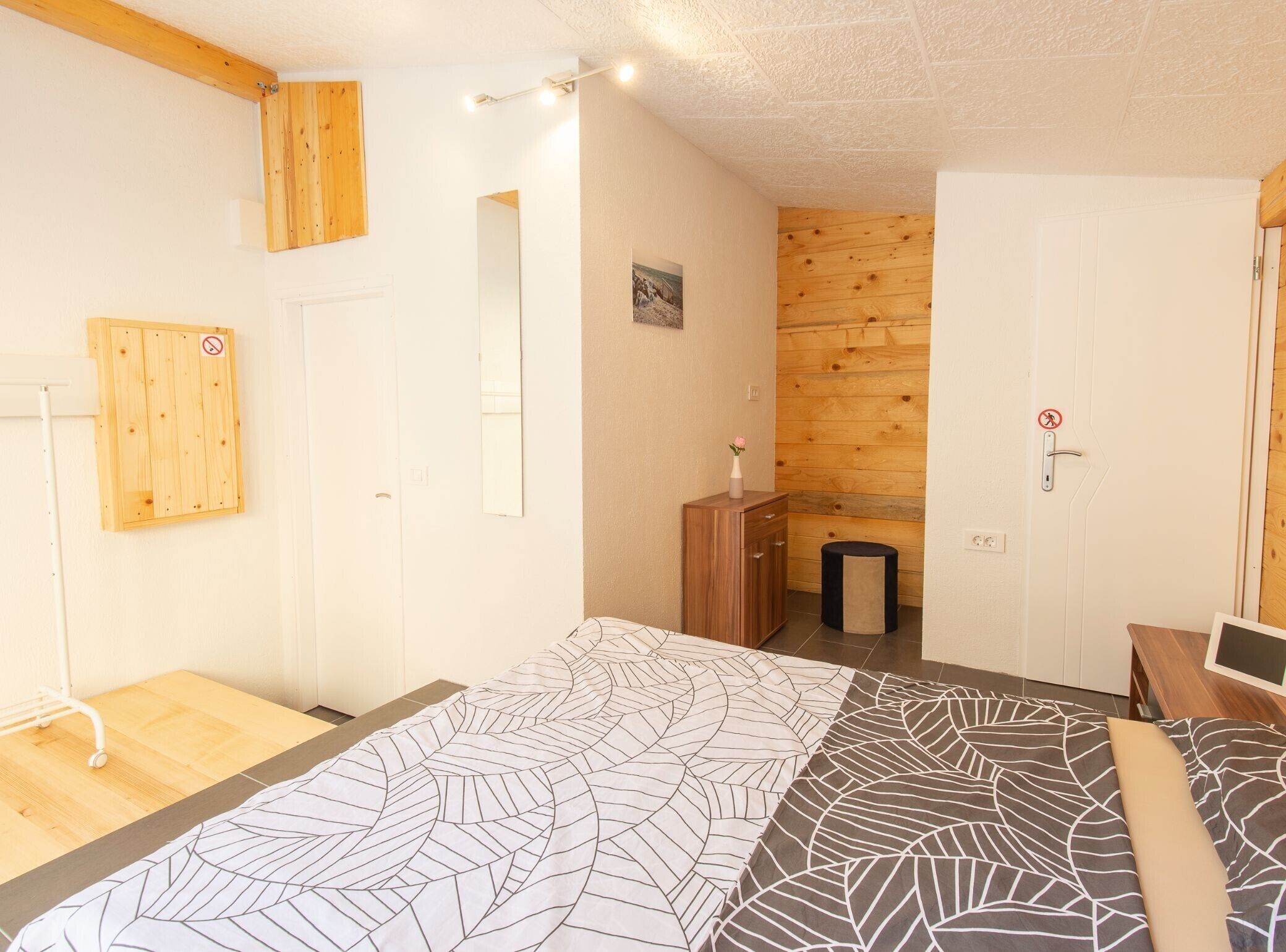 A bedroom with a bed, wooden walls, a small wooden cabinet, a mirror, and a door with a no-smoking sign.