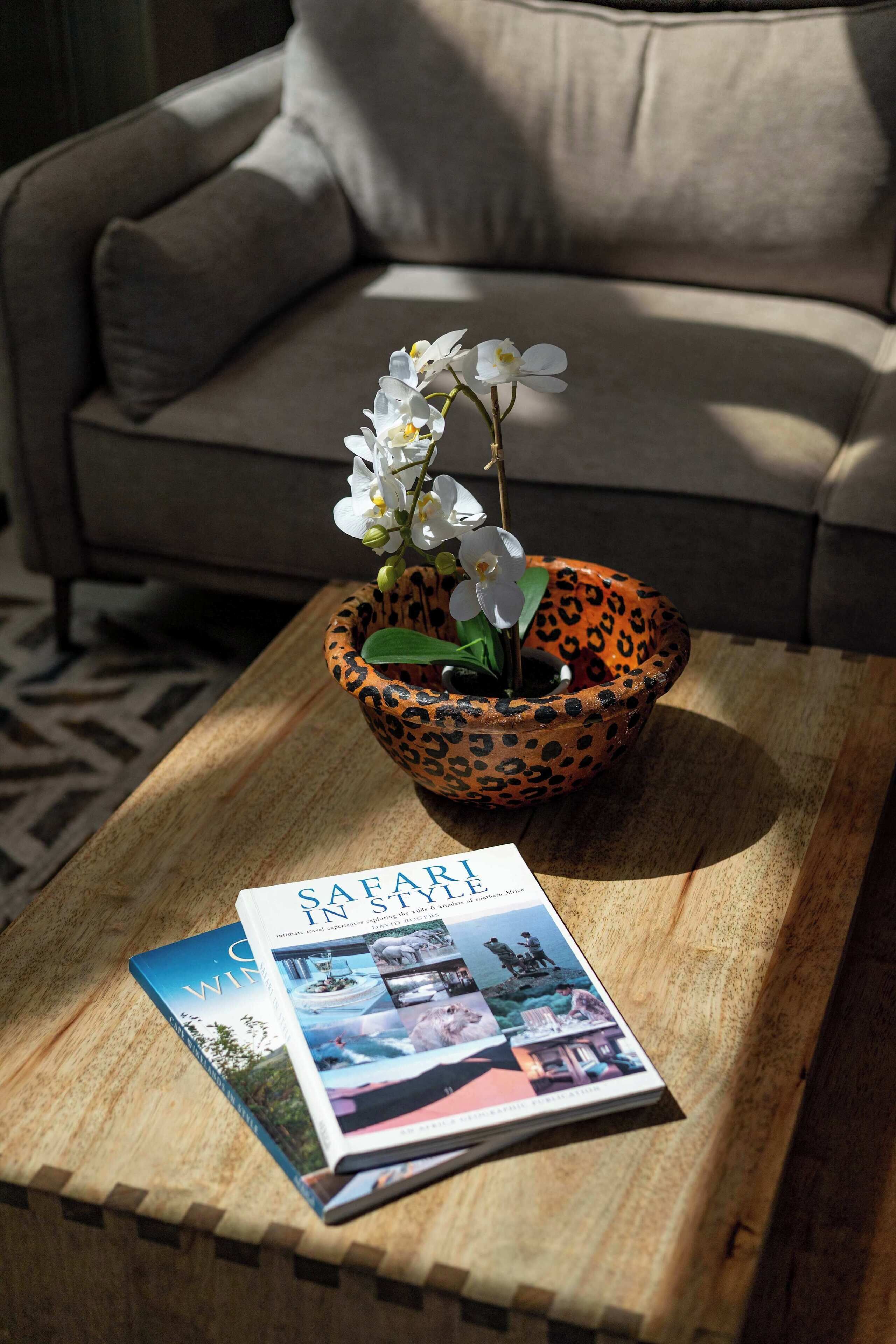 Eine Schale mit Leopardenmuster, darauf weiße Orchideen, auf einem Holztisch. Daneben ein Magazin mit der Aufschrift 'SAFARI IN THE SOUTH' und im Hintergrund ein beiges Sofa.