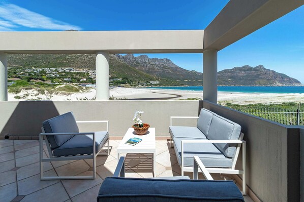 A balcony with two grey lounge chairs, a small table with a bowl of flowers, and a view of a beach and mountains.
