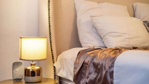 Een net opgemaakt bed met witte lakens en een bruine plaid, een bedlamp met gouden voet en een klein, gestructureerd object op het nachtkastje.