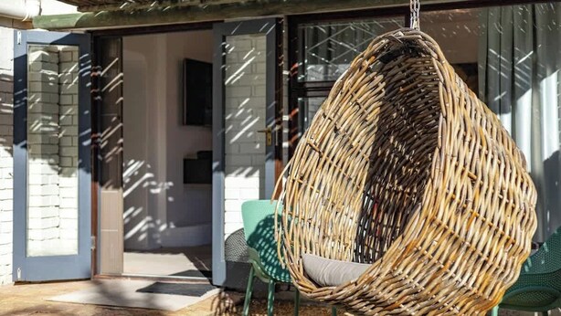 Een hangstoel van riet met een kussen, groene stoelen en een glazen deur die naar een binnenruimte leidt.