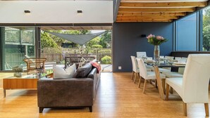 A modern living room with a sofa, dining table, and chairs. There is a large window providing natural light and a view of the outdoors. - Luxury Eco Haven With Garden Cottage - Hout Bay (Cape Town)