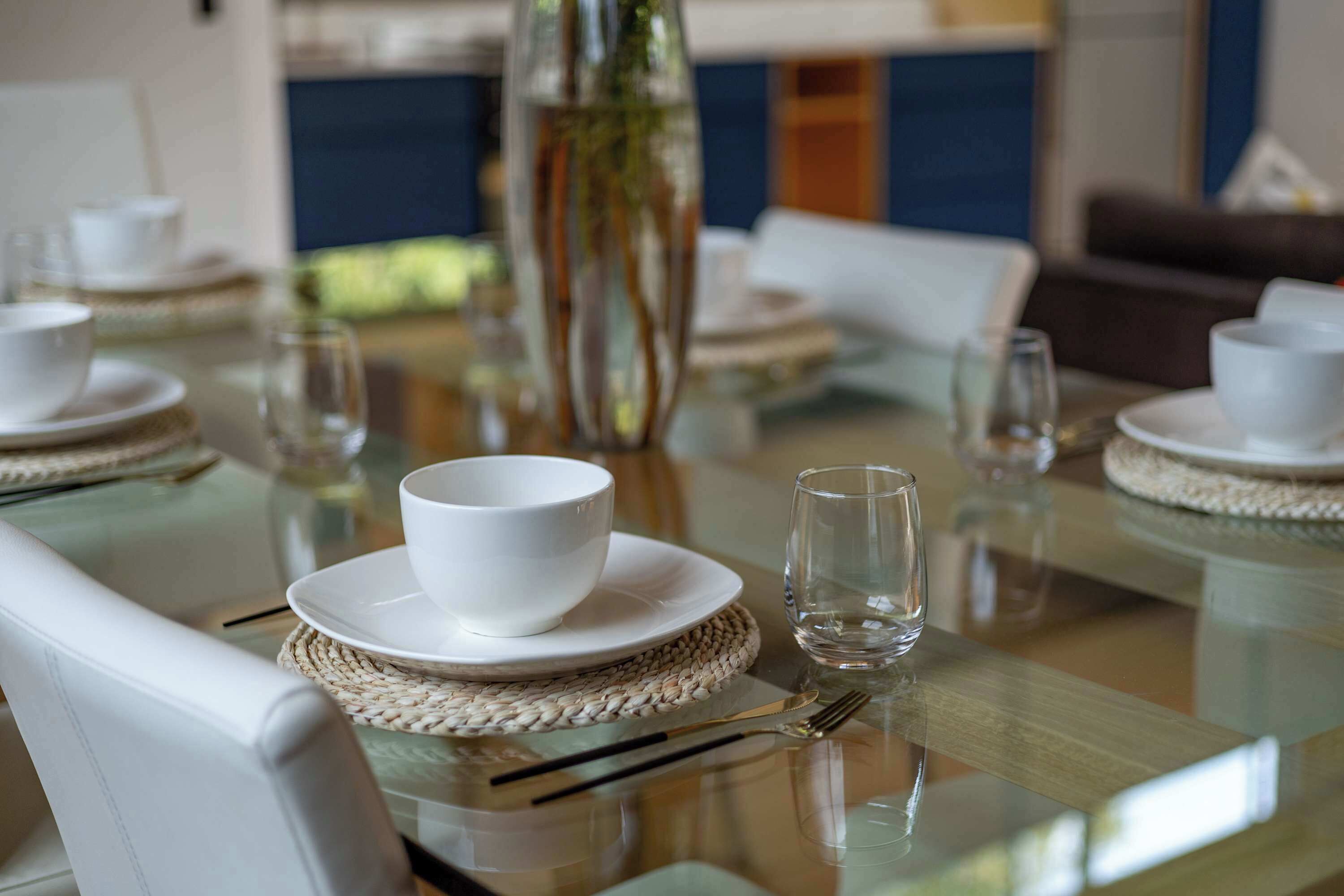 Una mesa de comedor dispuesta con platos, tazones y vasos blancos, con individuales tejidos y una cubierta de vidrio transparente.