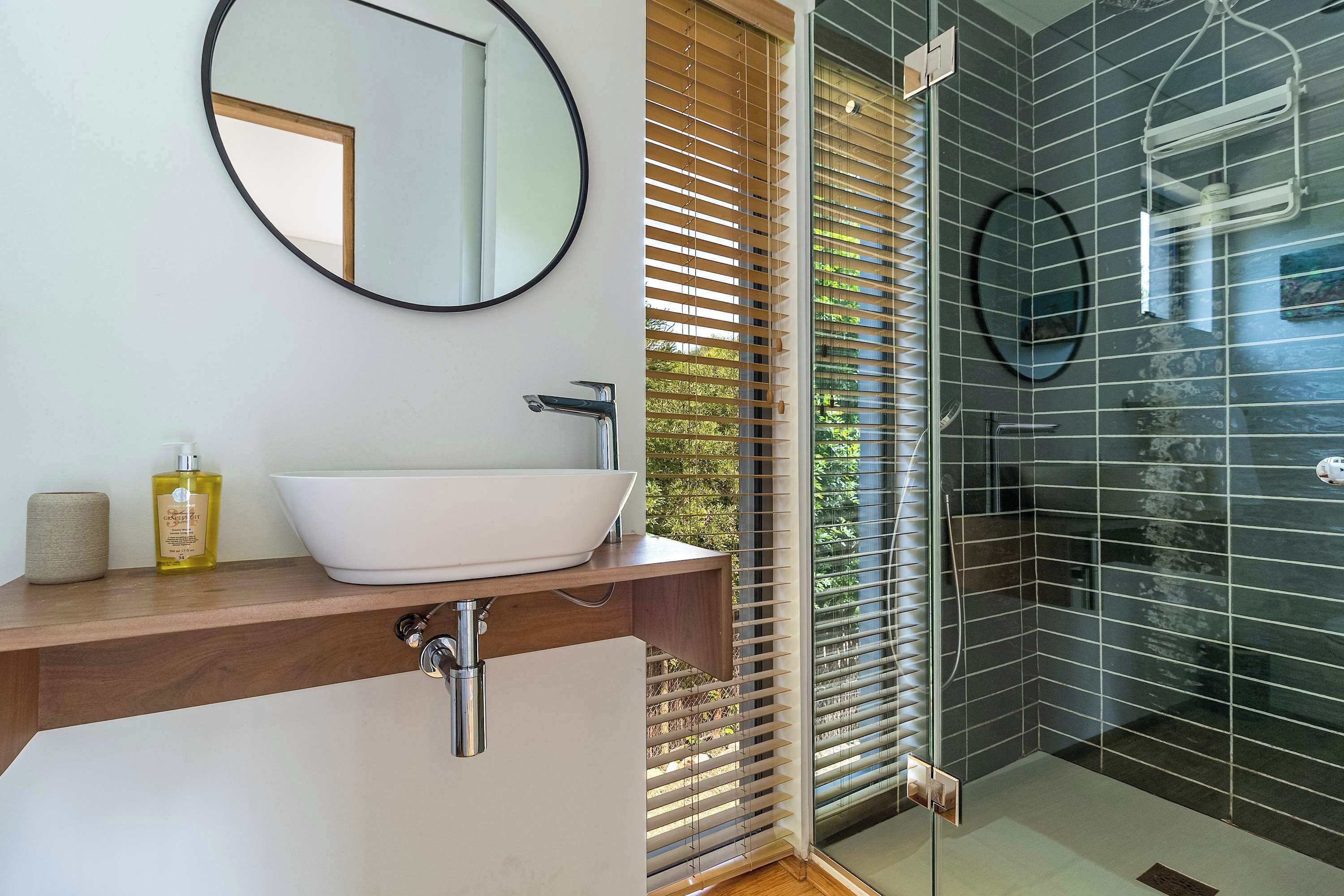 A bathroom with a white vessel sink, wooden vanity, a round mirror, and a glass-enclosed shower area with a black tiled wall.