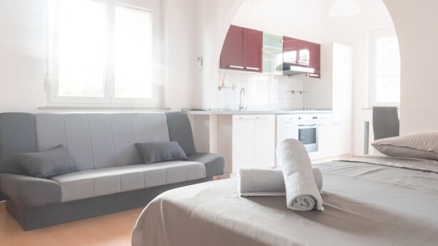Ein modernes, helles Zimmer mit einem grauen Sofa, einem ordentlich bezogenen Bett mit einem aufgerollten Handtuch und einer durch einen Bogen sichtbaren Küchenzeile.