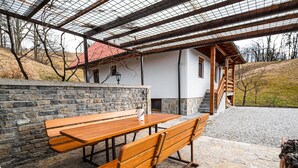 Une terrasse en pierre avec des bancs en bois et une table, un espace extérieur couvert avec un toit en métal, et un bâtiment avec un toit rouge en arrière-plan.