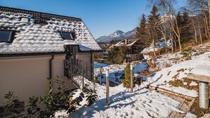 Una casa cubierta de nieve con un balcón, una montaña al fondo y un cielo azul despejado.