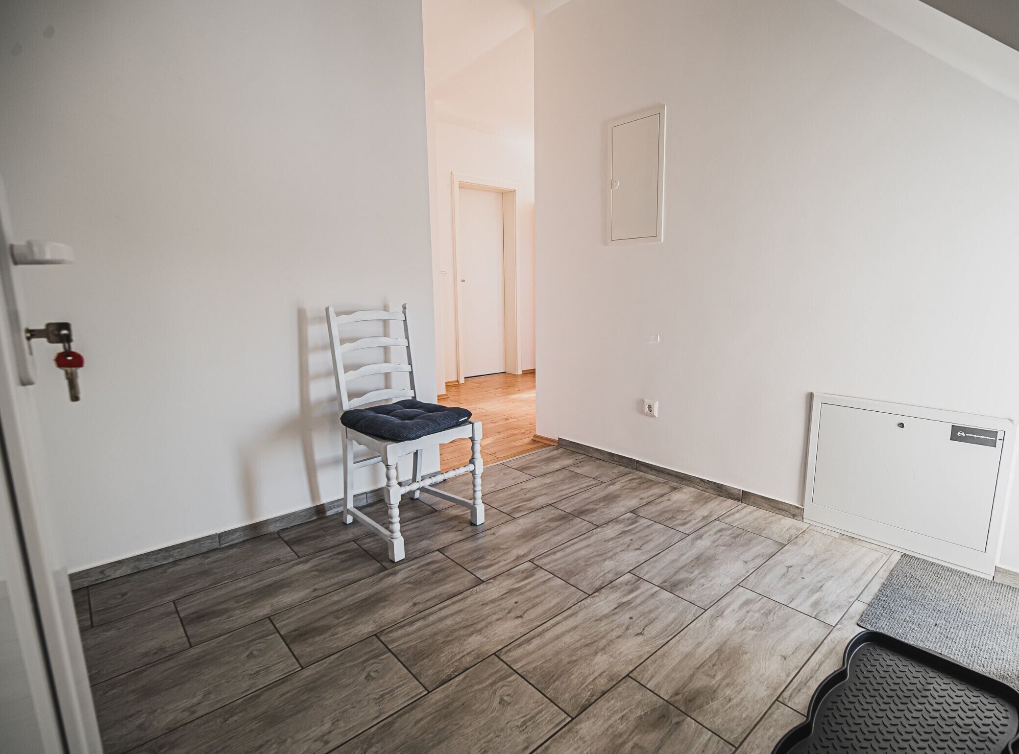 Une pièce avec une chaise blanche, un plancher en bois, un radiateur et une porte dotée d’une serrure à clé.