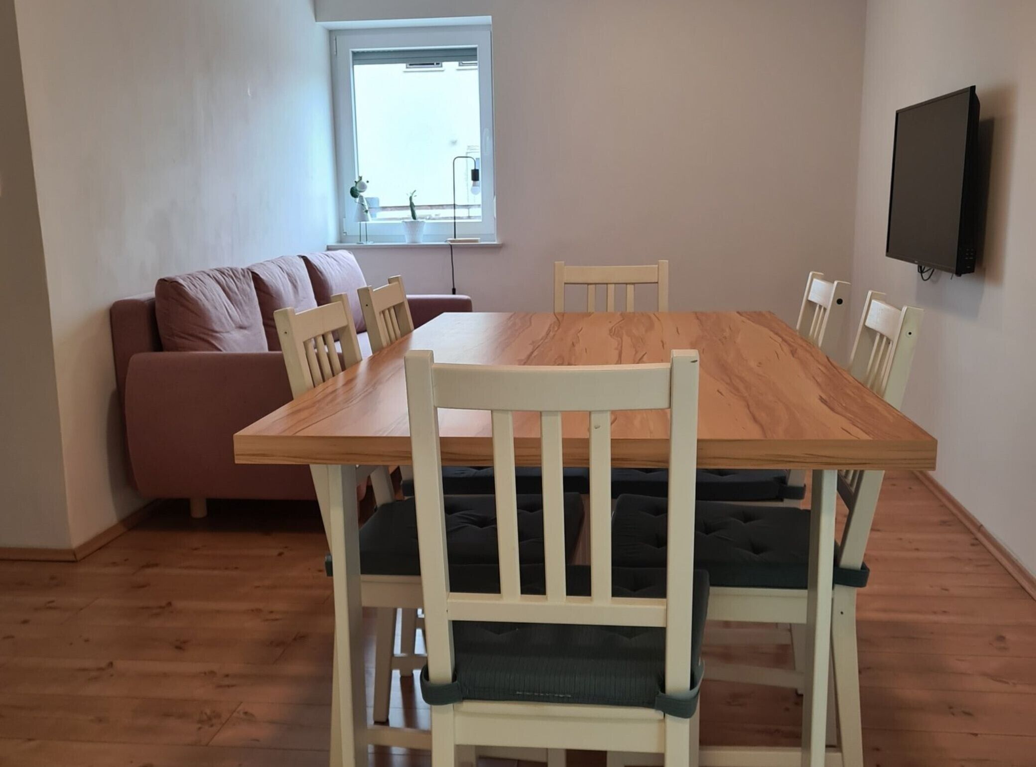 Un comedor con una mesa de madera y sillas blancas, un sofá y un televisor instalado en la pared.