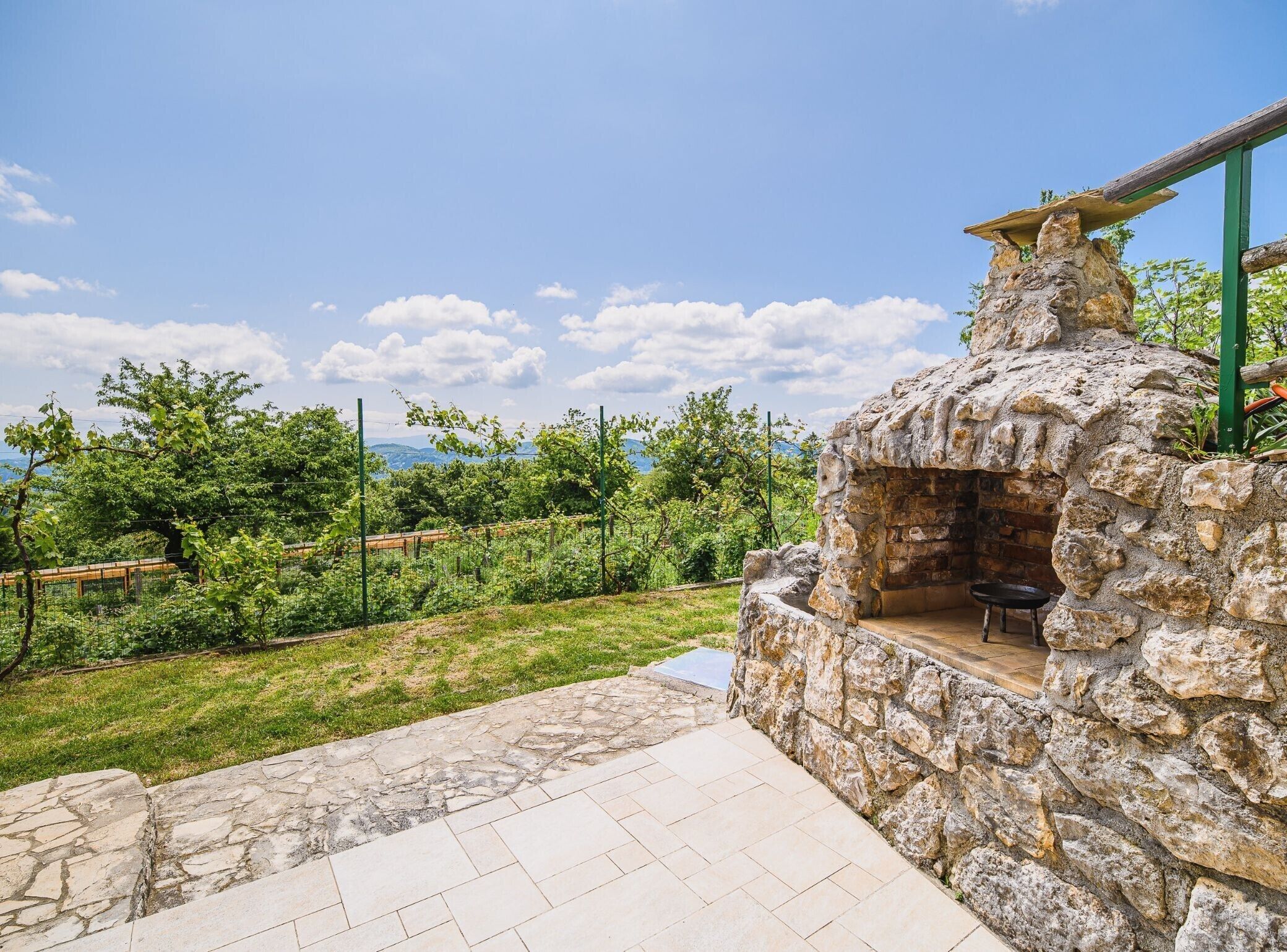 A stone-built outdoor kitchen with a stone bench, surrounded by a grassy area and a vineyard.