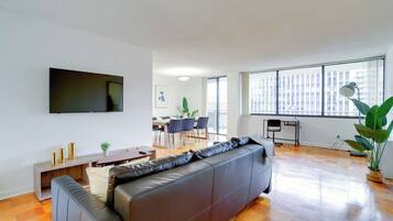 Ein modernes Wohnzimmer mit einer Ledersofa, einem Flachbildfernseher an der Wand, einem Essbereich mit Stühlen und einem Tisch sowie großen Fenstern mit Blick auf die Stadt.