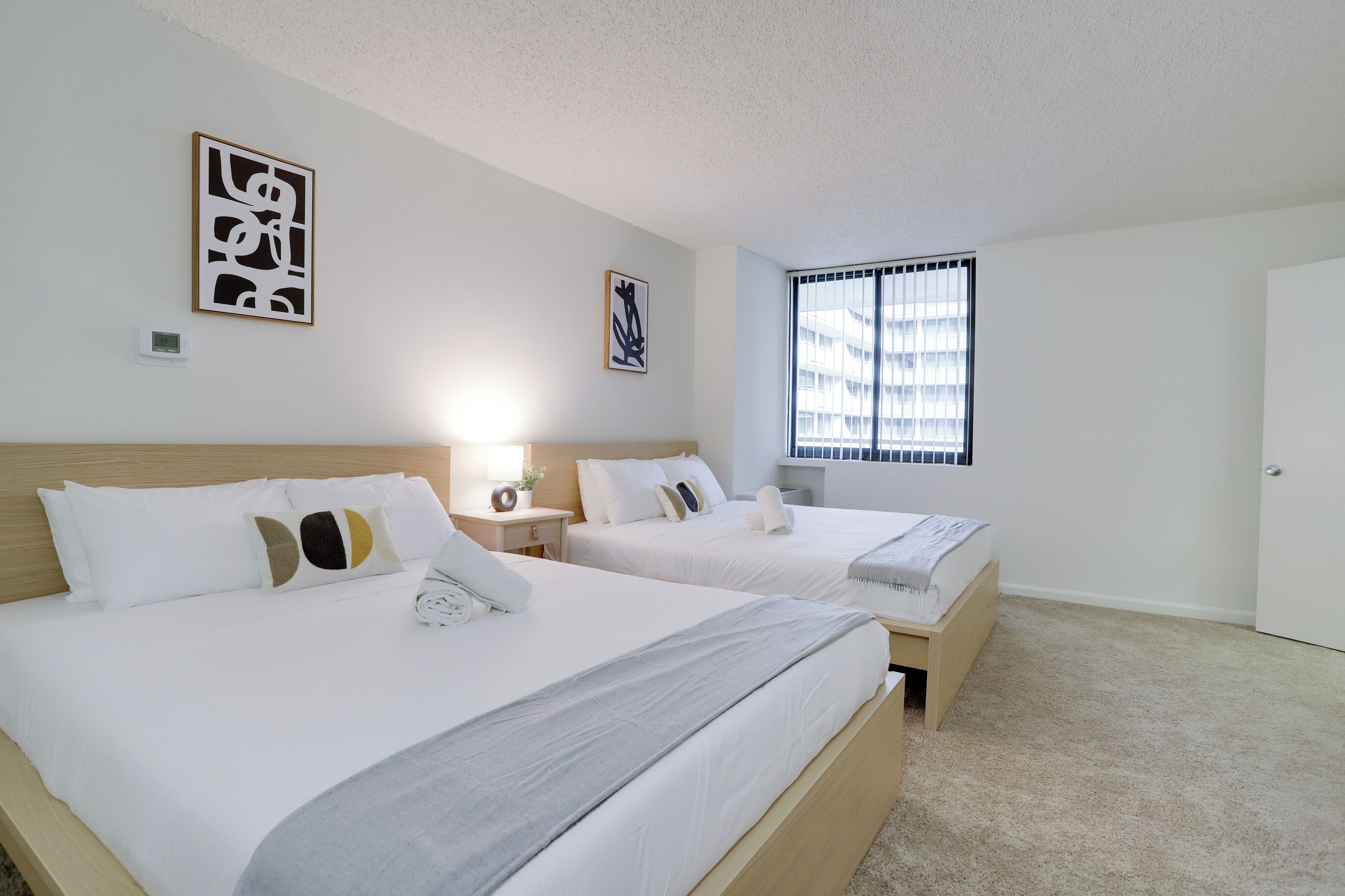 A hotel room with two beds, a window with blinds, and artwork on the walls.