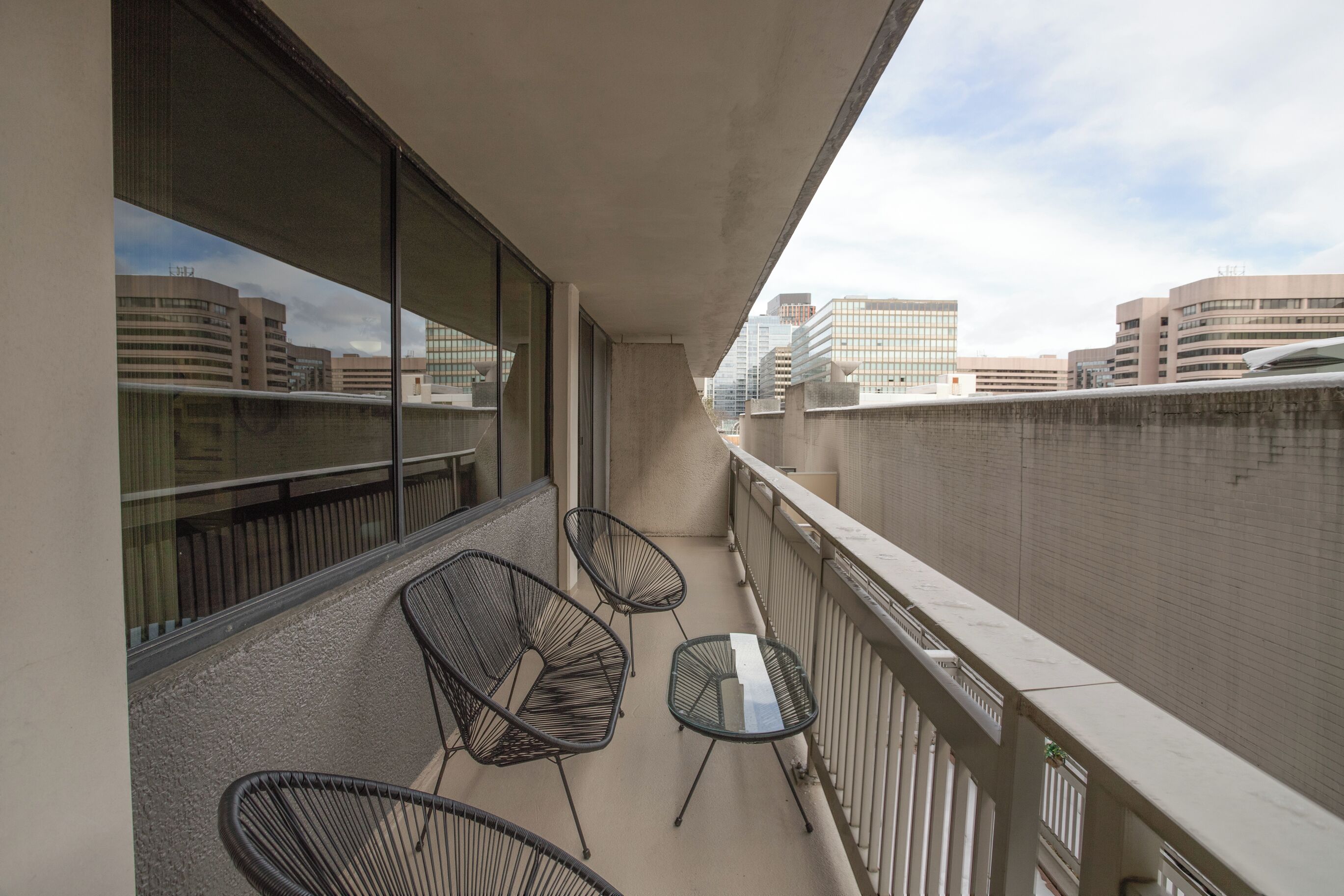 Varanda com duas cadeiras de arame pretas e uma pequena mesa, com vista para um horizonte urbano com edifícios modernos.