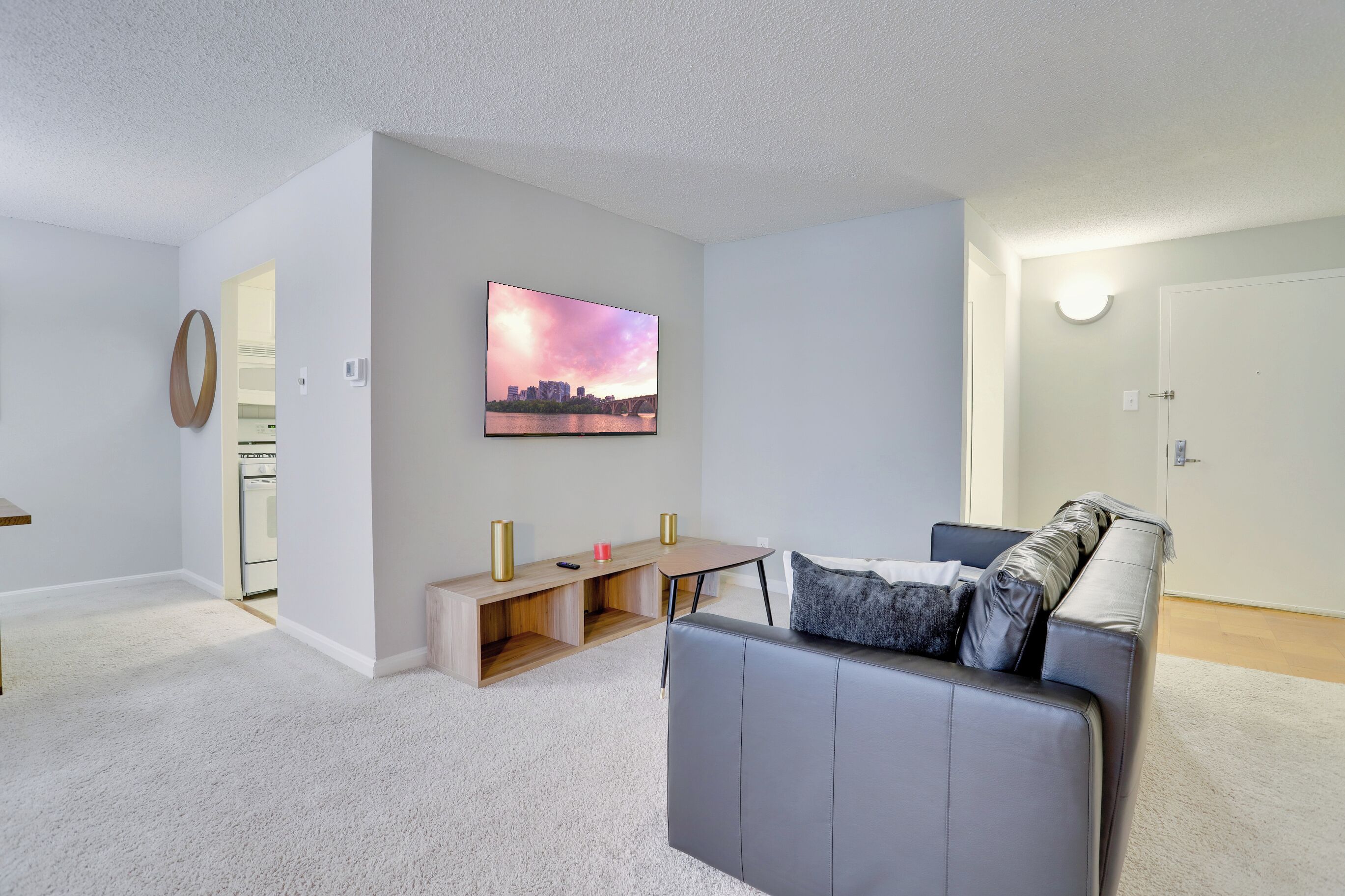 A modern living room with a grey sofa, a coffee table, and a painting of a cityscape. There is a wooden desk with two gold lamps and a small pink object on it. The room has white walls and a carpeted floor.