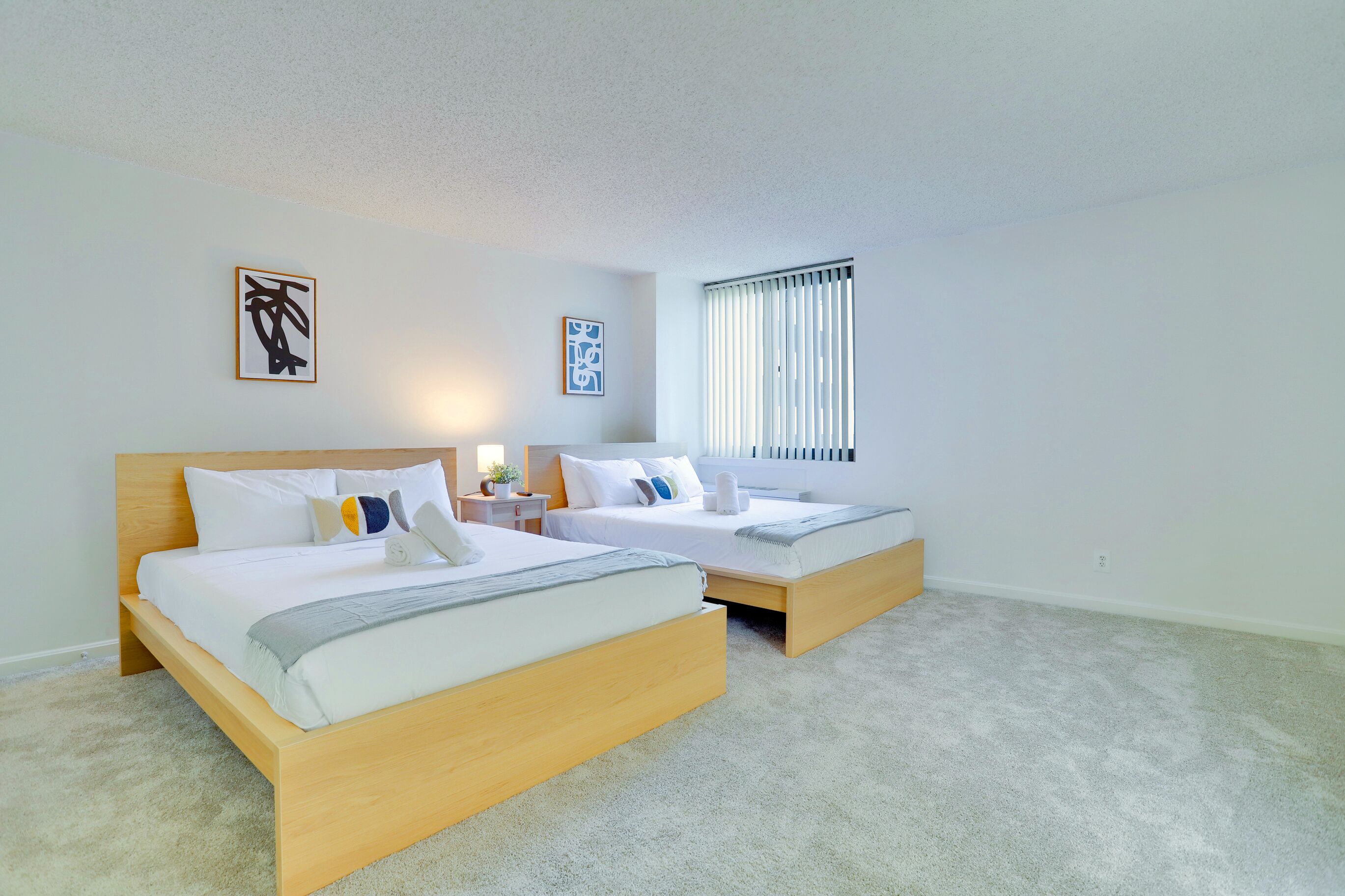 A hotel room with two single beds, a nightstand, a lamp, and a window with blinds.