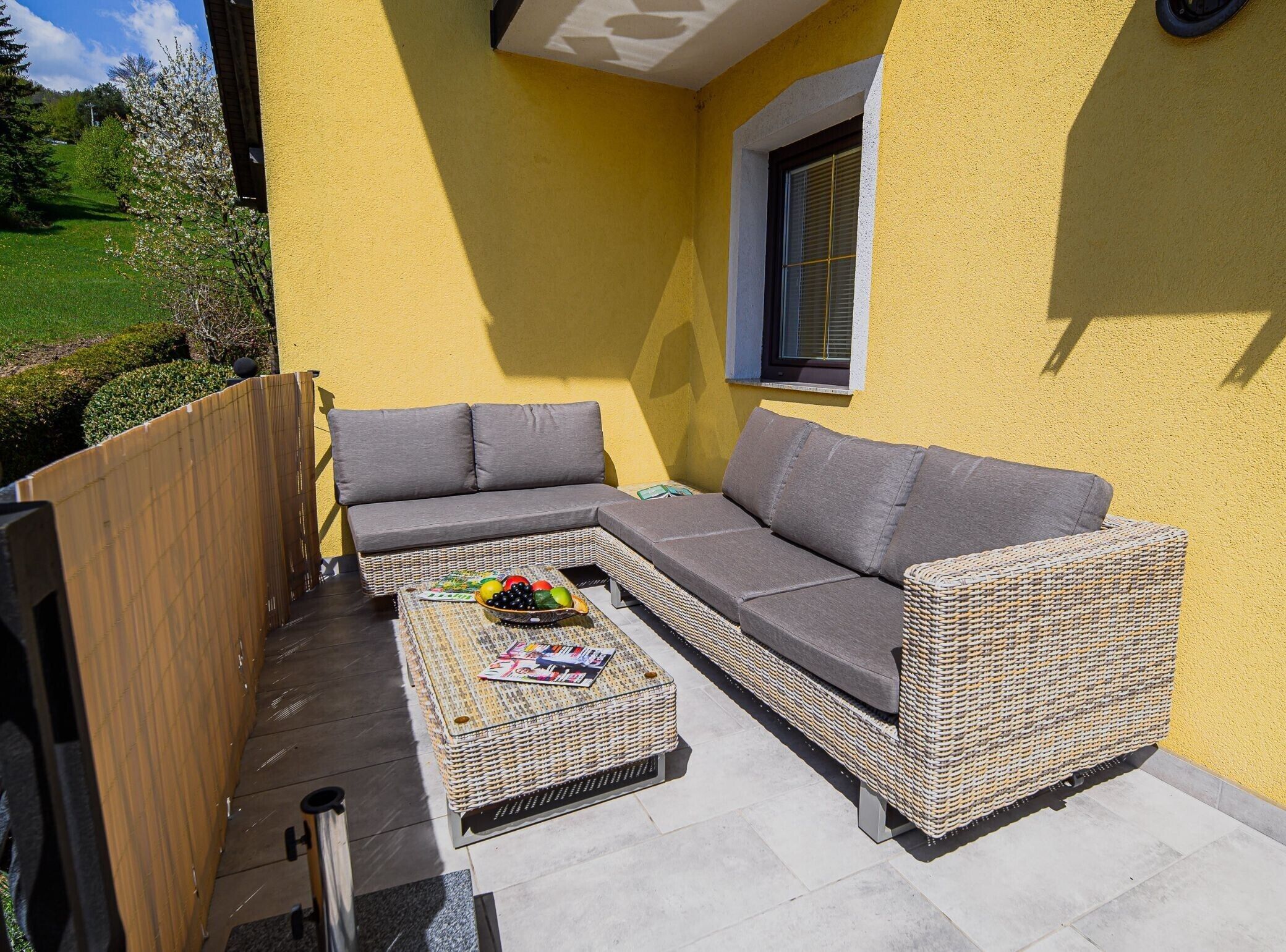 A yellow exterior wall with a window, a wicker sofa, and a small table with magazines and fruit.