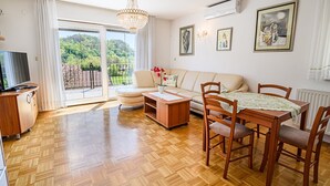 Un salon spacieux avec un canapé, une table basse en bois et une table à manger avec des chaises. On y trouve un téléviseur posé sur un meuble en bois, et un lustre est suspendu au plafond.