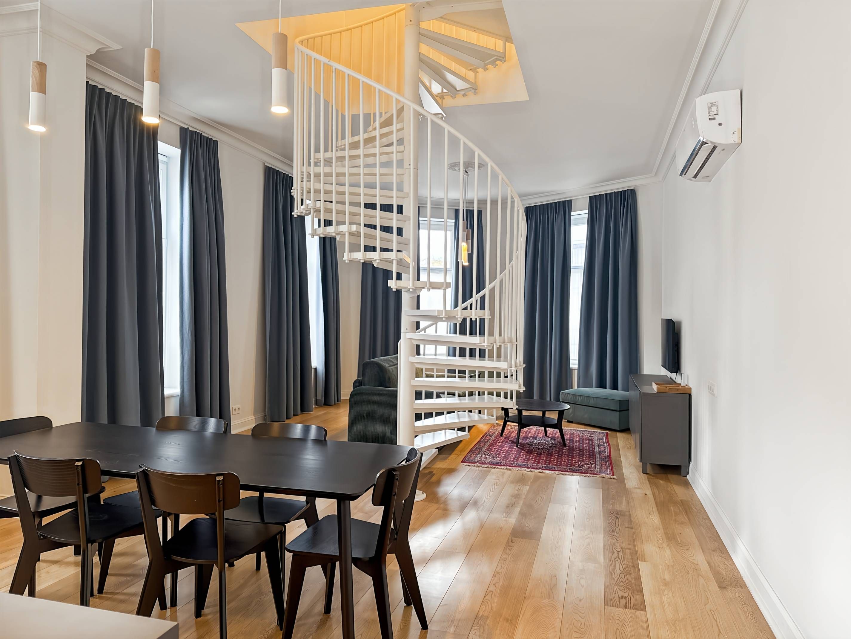 Un salon moderne avec un escalier en colimaçon, un coin repas avec une table et des chaises en bois, et un canapé gris.