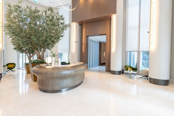 Un hall d’entrée moderne avec un comptoir d’accueil central, un grand arbre intérieur et un espace lumineux et vaste offrant une vue sur la ville.