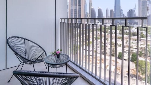 Un balcon avec deux chaises à l’assise en fil de fer noir et une petite table ronde agrémentée d’une fleur. Le balcon offre une vue sur un paysage urbain avec de hauts immeubles.