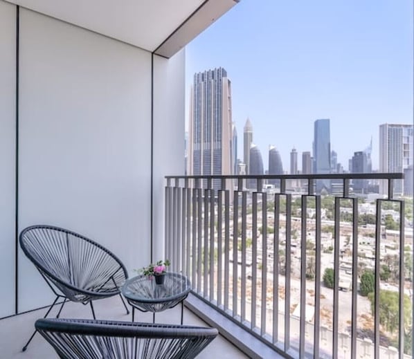 Ein Balkon mit zwei schwarzen Stühlen aus Draht und einem kleinen runden Tisch mit einer Blume. Der Balkon bietet Blick auf eine Skyline mit hohen Gebäuden.