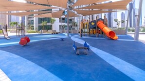 Een speelplaats met een glijbaan en klimconstructies, omringd door een blauwe vloer en beschaduwd door grote luifels.