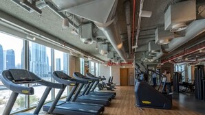 Een sportschool met loopbanden, een krachttrainingapparaat en een uitzicht op de stadshemel door grote ramen.