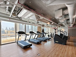 A modern gym with treadmills and elliptical machines, large windows offering city views, and exposed ductwork on the ceiling.
