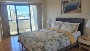 A bedroom with a bed, a nightstand, a lamp, a view of the beach, and a balcony. - Holiday In Husky by Book Jervis Bay - 2 Bedroom Apartment located in the centre of town (Huskisson)