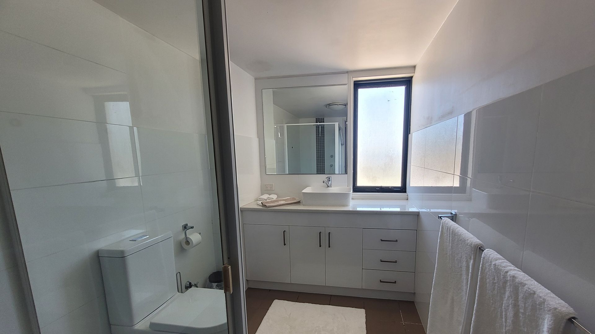 A modern bathroom with a glass shower enclosure, a toilet, a sink with a mirror, and a window providing natural light.