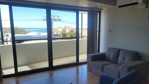 A room with a large sliding glass door leading to a balcony with a view of the sea and buildings. There is a grey sofa in the room. - Holiday In Husky by Book Jervis Bay - 2 Bedroom Apartment located in the centre of town (Huskisson)
