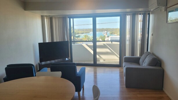 A modern living room with a flat-screen TV, a grey sofa, a wooden dining table, and a large sliding glass door leading to a balcony with a view of a marina and hills. - Holiday In Husky by Book Jervis Bay - 2 Bedroom Apartment located in the centre of town (Huskisson)