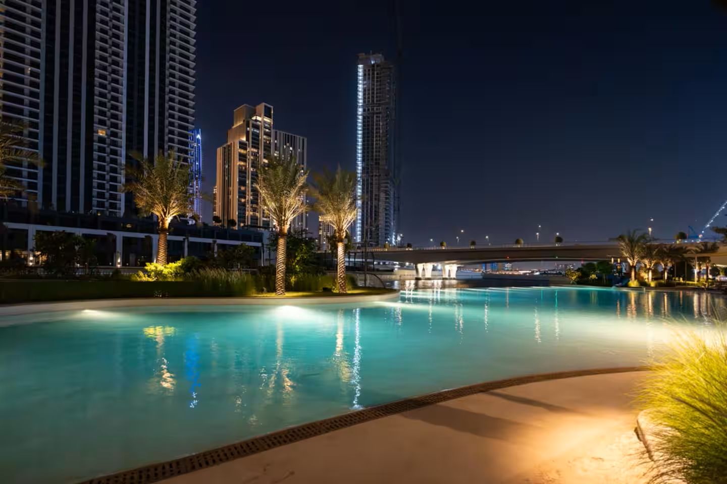 Ein Pool bei Nacht mit umliegenden Palmen und Hochhäusern im Hintergrund.