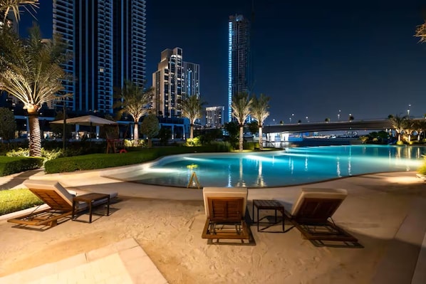 A nighttime pool area with lounge chairs, a well-lit pool, and high-rise buildings in the background.