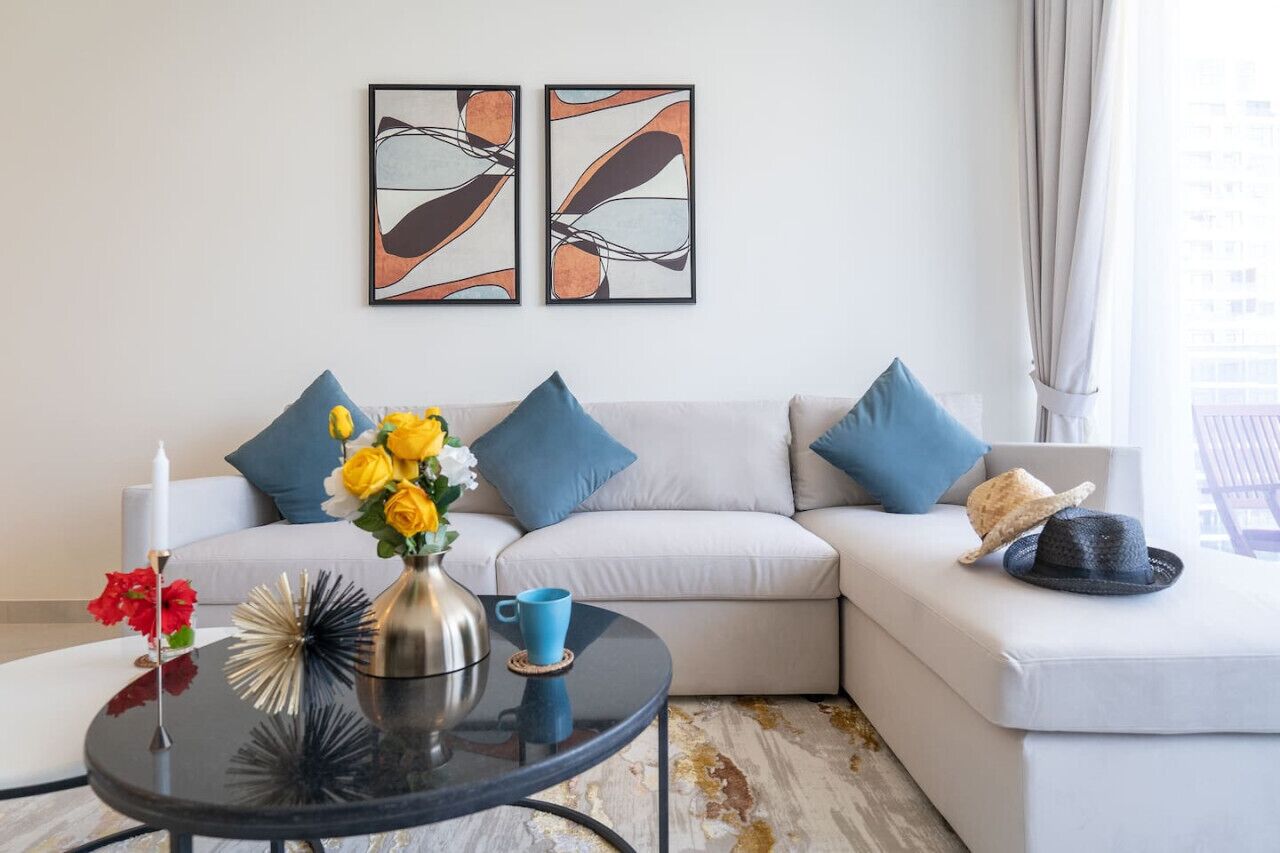 Una sala moderna con un sofá de color claro, cojines azules, una mesa de centro redonda con un jarrón de flores amarillas y arte abstracto en la pared.