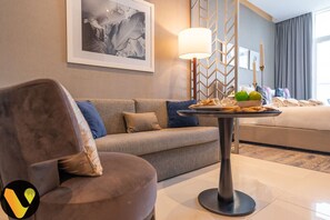 A modern hotel room with a sofa, a table with fruits and towels, a lamp, and a framed picture on the wall.