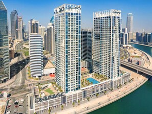A high-rise building with a prominent 'DAMAC' sign, surrounded by other modern high-rises, a waterfront, and a busy road.