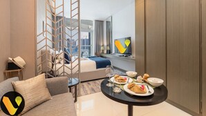 A modern hotel room with a bed, a sofa, a dining table with breakfast, and a view of the city through large windows.