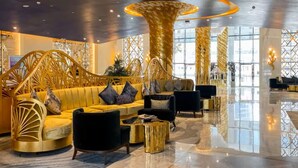 A spacious lobby with a modern design, featuring a central seating area with a sofa and armchairs, a large decorative column, and a reflective floor.