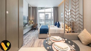 A modern hotel room with a bed, a sofa, a dining table set for two, a TV, and a city view.