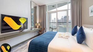 A hotel room with a large bed, a flat-screen TV, a desk with a lamp, and a view of the city through large windows.