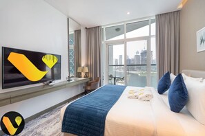 Habitación de hotel con una cama grande, un televisor de pantalla plana, un escritorio con lámpara y vistas a la ciudad a través de amplias ventanas.