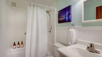 A bathroom with a shower, toilet, sink, and a mirror. There is a towel hanging on the wall and three bottles on the floor.