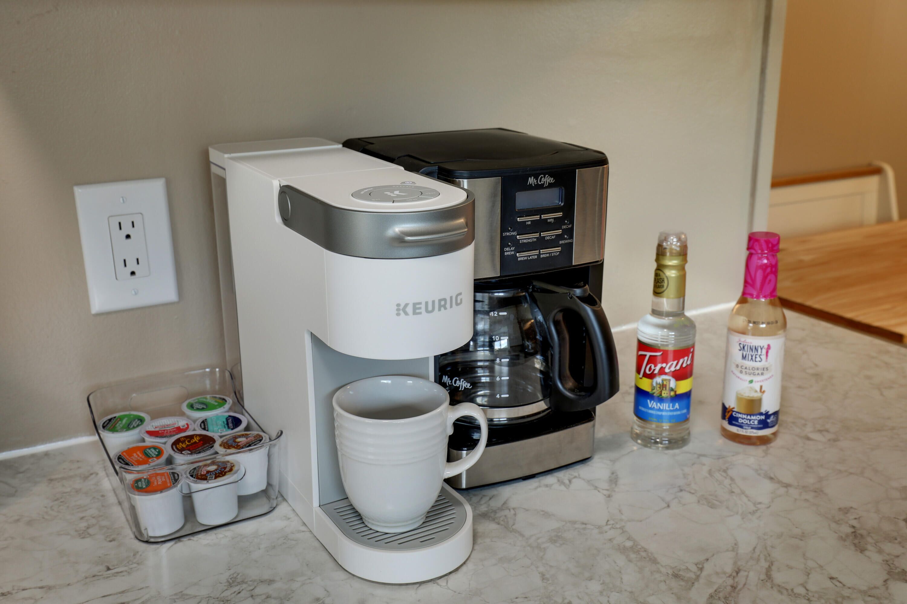 A Keurig coffee maker with a mug, a coffee pod holder, and two bottles of Torani syrup on a kitchen counter.