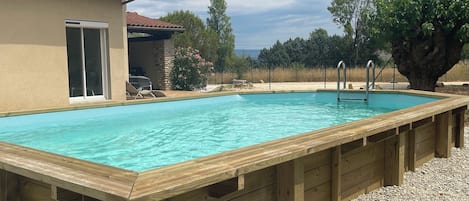 En rektangulær swimmingpool med en træterrasse hele vejen rundt, en bygning med stenvægge og et tag med fliser, og et stort træ i baggrunden.