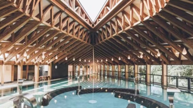 A large indoor swimming pool with a unique wooden ceiling structure and a central viewing platform.