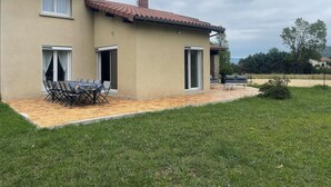 A two-story house with a tiled roof, a patio with a table and chairs, and a grassy yard.