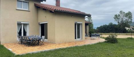 A two-story house with a tiled roof, a patio with a table and chairs, and a grassy yard.