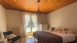 A bedroom with a wooden ceiling, a bed with a brown bedspread, a chair, a bedside table, and a window with curtains.