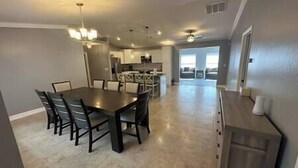 A spacious dining area with a large table and chairs, a kitchen in the background, and a view of the outdoors.